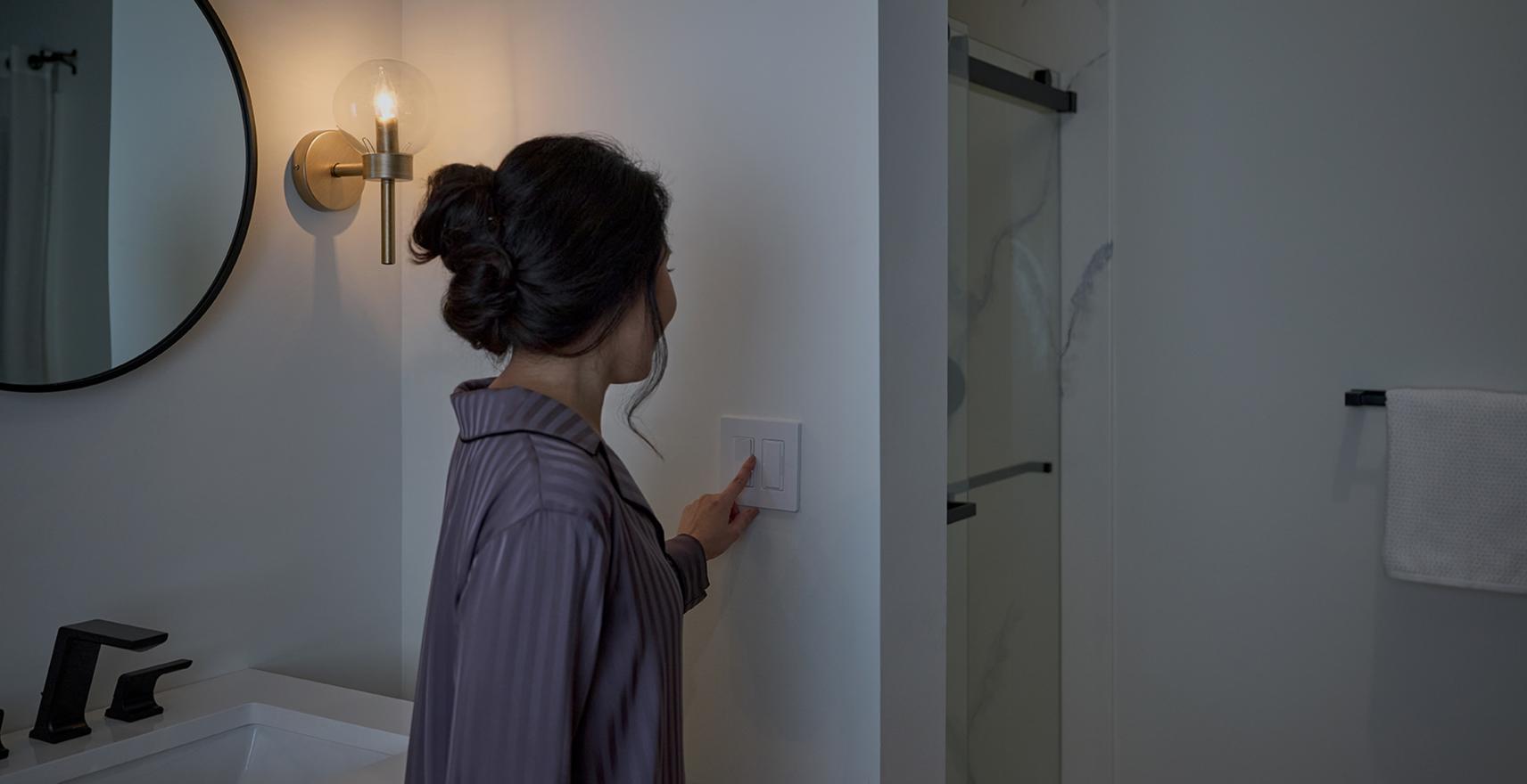Woman adjusting a Diva dimmer with Night Mode, ganged with a Humidity Sensor switch, in a bathroom setting.